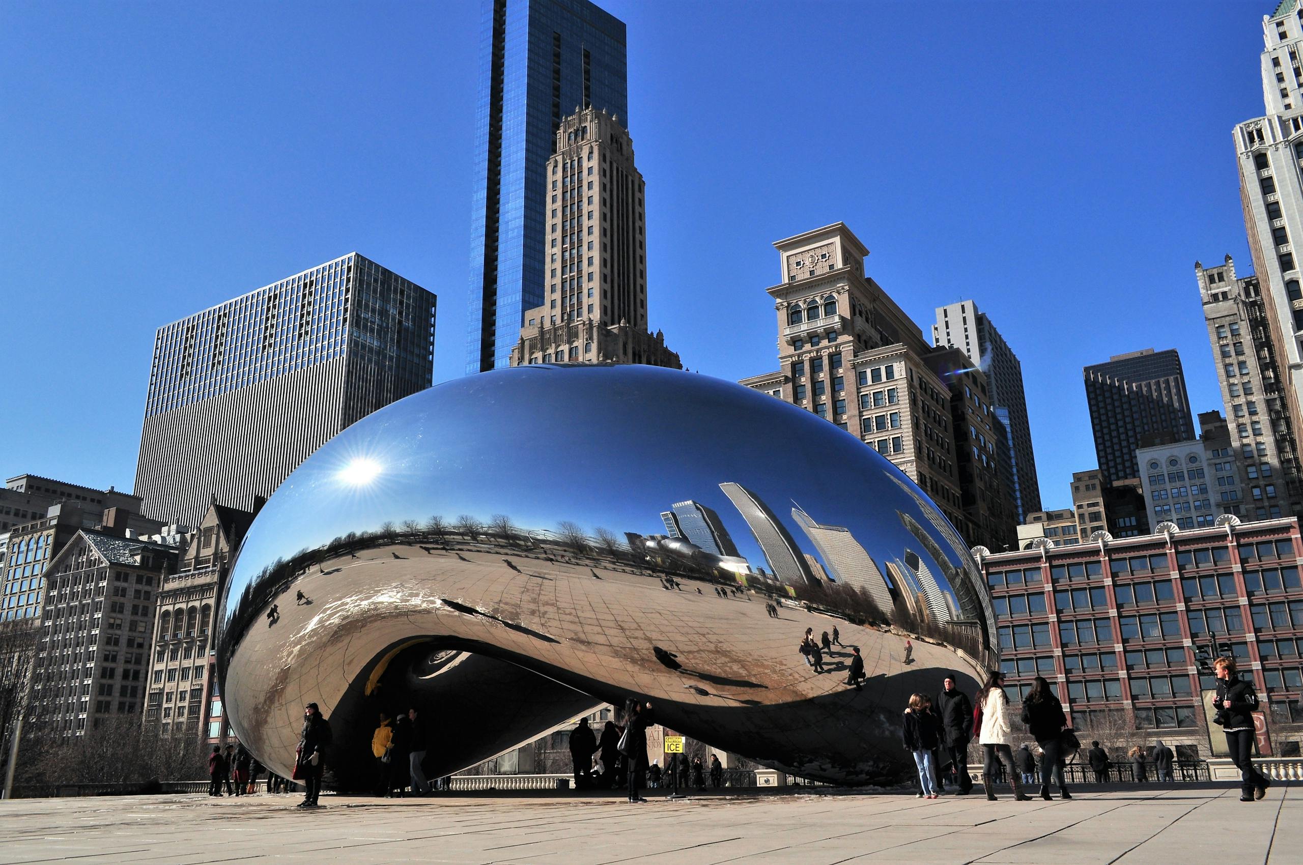 Reflective Cloud Gate sculpture in bustling Chicago cityscape with skyscrapers under a clear blue sky. Chicago hotel group rates. 